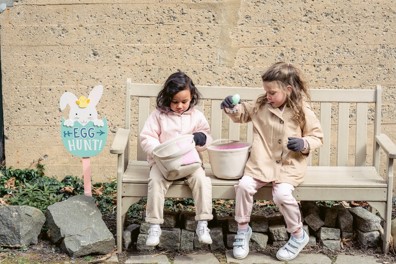 Two girls sharing easter eggs on a bench after an easter egg hunt, with an easter egg hunt sign next to the bench